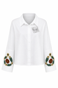 A white long-sleeve button-up shirt with a pointed collar and detailed floral embroidery on the cuffs, featuring a small silver brand tag on the chest. The brand name 'EVIA' is visible on the tag.