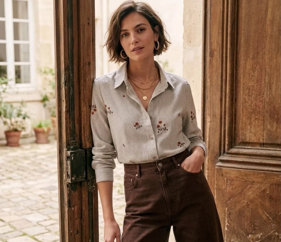 A woman wearing a light beige long-sleeve button-up shirt with delicate floral embroidery and dark brown trousers, standing in a rustic doorway.