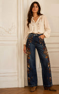 A woman stands indoors against a white paneled wall wearing wide-leg blue jeans with ornate multicolor floral embroidery, a cream embroidered blouse, and brown ankle boots.