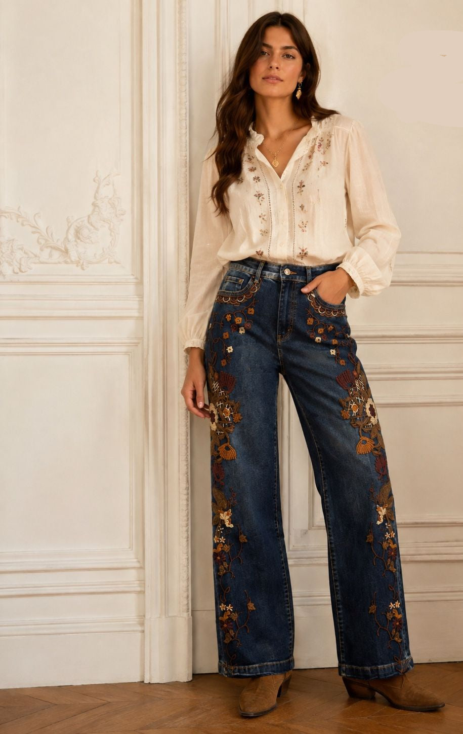 A woman stands indoors against a white paneled wall wearing wide-leg blue jeans with ornate multicolor floral embroidery, a cream embroidered blouse, and brown ankle boots.