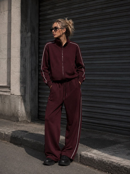 A woman wearing a matching burgundy zip-up jacket and wide-leg sweatpants with white side stripes, standing outdoors in front of a dark metal shutter.