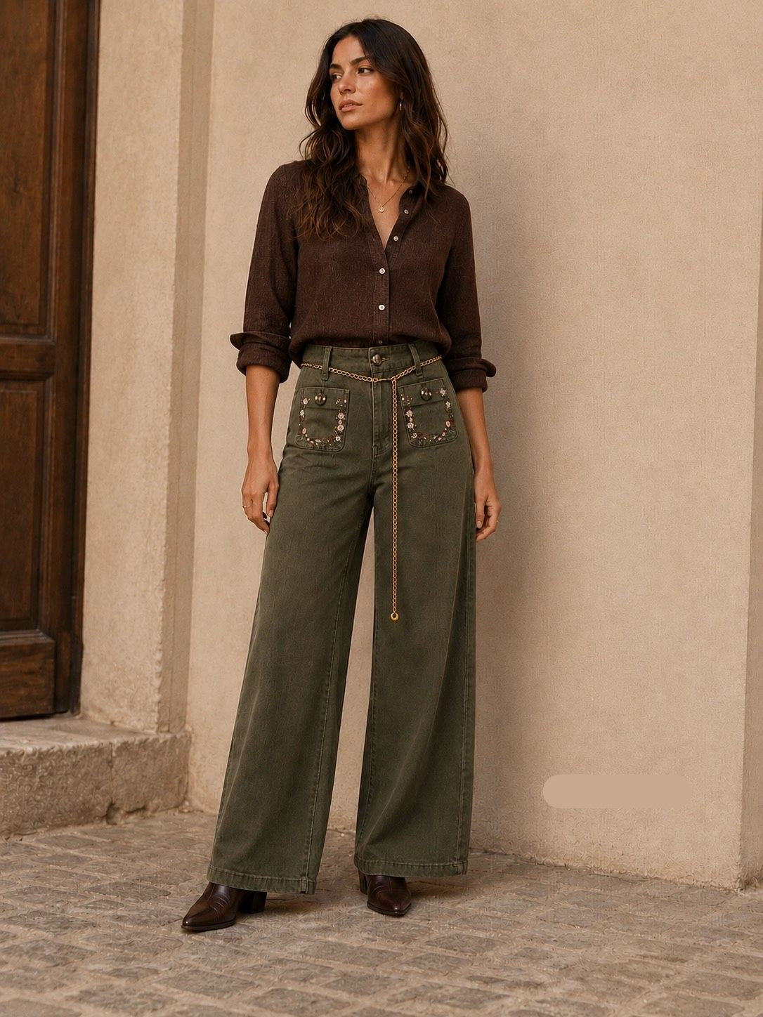 A woman stands outdoors against a beige stucco wall wearing a dark brown button-front shirt and olive green wide-leg pants with decorative pocket details and a long tie belt. She is also wearing dark brown shoes.