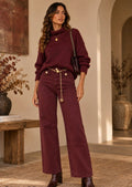A woman models a matching burgundy sweater and wide-leg jeans set with gold chain and belt details, standing in a warm, rustic interior setting.
