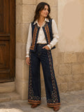 A woman stands outdoors against a stone wall wearing a white button-up shirt, a dark blue embroidered vest, wide-leg dark blue jeans with ornate embroidery down the sides and at the hems, and brown boots. Text on the right reads 'Automne mi saison' with garment codes.