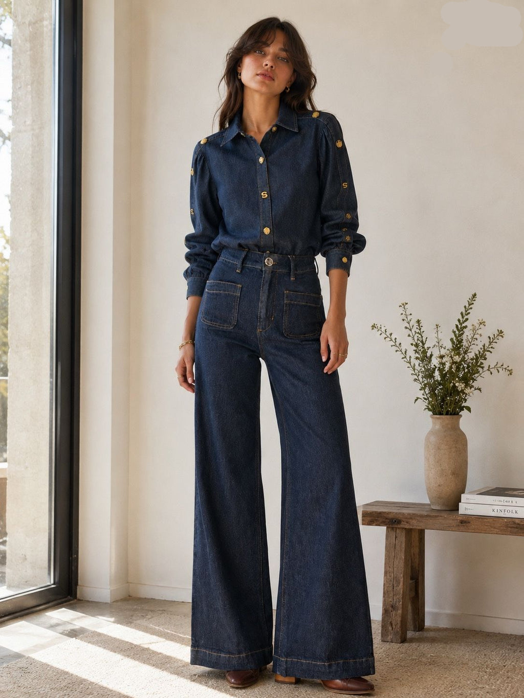 A woman models a dark blue denim two-piece set consisting of a long-sleeve button-up shirt with gold buttons and matching high-waisted wide-leg jeans, standing in a brightly lit room next to a window.