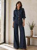 A woman models a dark blue denim two-piece set consisting of a long-sleeve button-up shirt with gold buttons and matching high-waisted wide-leg jeans, standing in a brightly lit room next to a window.