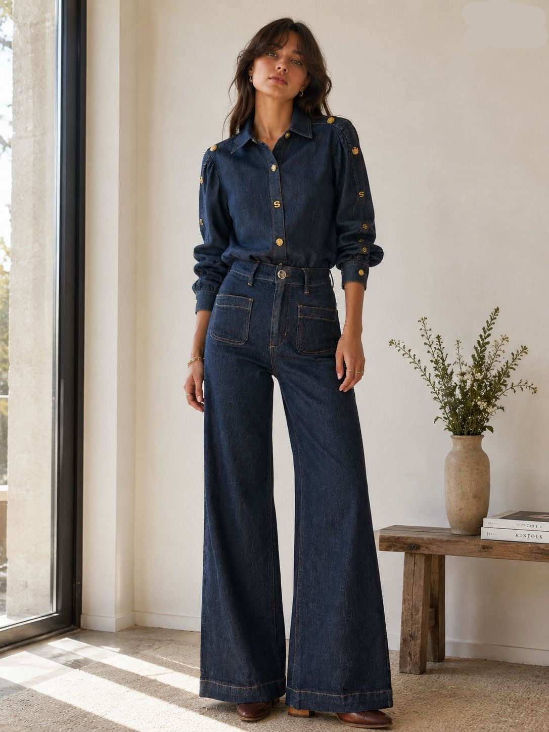 A woman models a dark blue denim two-piece set consisting of a long-sleeve button-up shirt with gold buttons and matching high-waisted wide-leg jeans, standing in a brightly lit room next to a window.