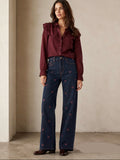 A woman stands indoors wearing a burgundy ruffled-sleeve blouse and dark blue wide-leg jeans with small embroidered heart motifs, paired with dark brown shoes. The background is a warm beige textured wall and floor.