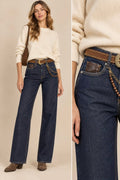 A model wears dark blue wide-leg jeans with a brown leather belt and chain detail, paired with a cream sweater and brown boots, shown in a full-body view and a close-up of the waist area.