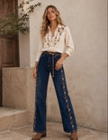A woman stands outdoors wearing a cream embroidered button-up shirt and dark blue wide-leg jeans with floral embroidery down the sides, paired with brown ankle boots. The background shows a textured beige stucco wall and stone steps.