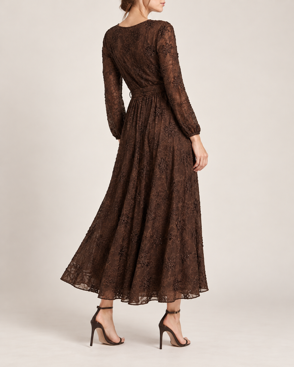 A woman modeling a long-sleeve brown lace maxi dress from the back, paired with black high-heeled sandals, against a plain light beige studio background.