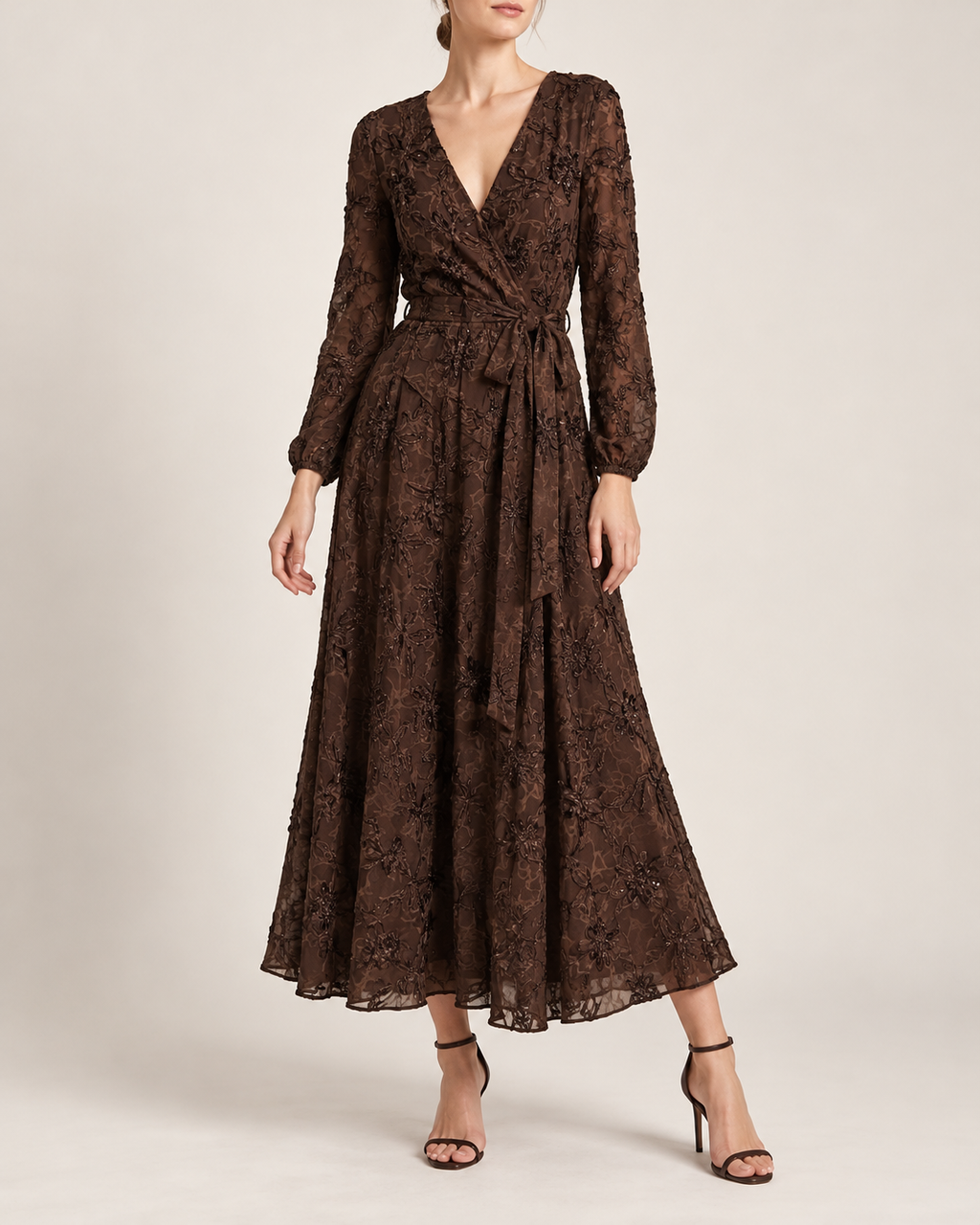 A woman models a long-sleeve, dark brown lace midi dress with a wrap-style V-neck and a matching fabric tie belt at the waist, paired with black strappy high-heeled sandals against a plain beige studio background.