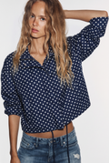 A woman wearing a navy blue long-sleeve button-up shirt with a white polka dot pattern, paired with blue jeans, against a plain white background.