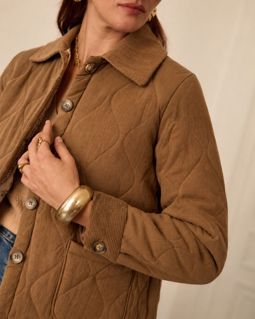 A close-up of a woman wearing a brown quilted jacket with a collar and large buttons, paired with a beige top and gold jewelry against a light neutral background.
