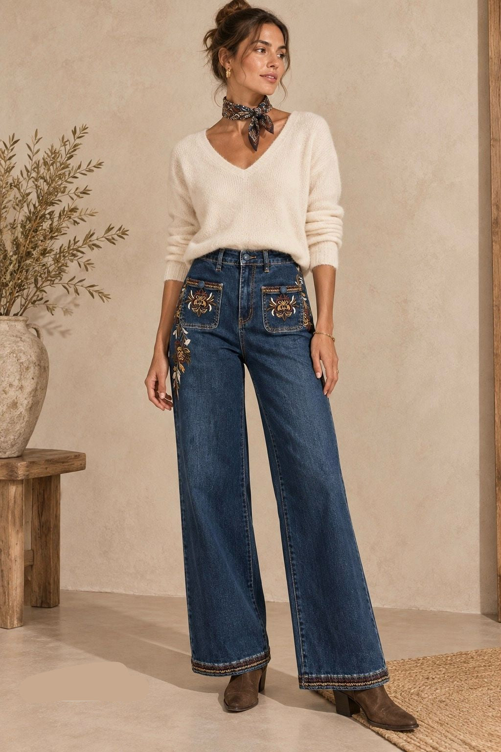 A woman models a pair of high-waisted, wide-leg blue jeans with decorative embroidered details on the front pockets, styled with a cream knit top and brown ankle boots in a warm, rustic interior setting.